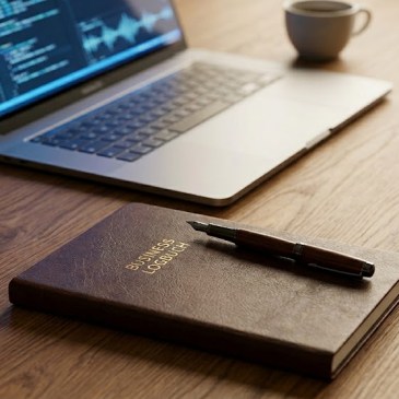 Ein minimalistischer, aufgeräumter Holzschreibtisch aus der Vogelperspektive oder leicht schräg von der Seite. Im Vordergrund liegt ein klassisches, haptisches Notizbuch mit einem hochwertigen Füllfederhalter. Im Hintergrund steht ein moderner Laptop, auf dessen Bildschirm schemenhaft Codezeilen oder digitale Wellenformen zu sehen sind.